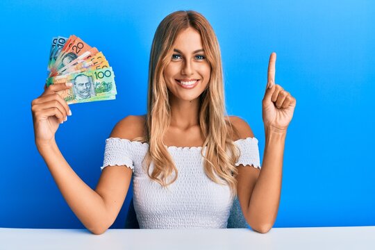 Beautiful blonde young woman holding australian dollars smiling with an idea or question pointing finger with happy face, number one