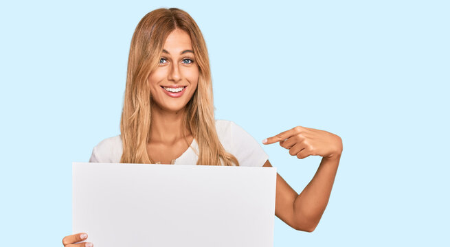 Beautiful blonde young woman holding blank empty banner pointing finger to one self smiling happy and proud