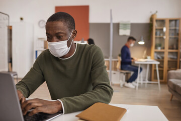 Portrait of African-American man wearing mask and using laptop while working at desk in office, copy space