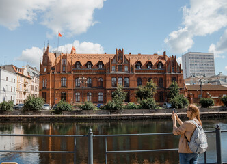 Young woman takes pictures of a beautiful building. House on the river. The girl with a backpack...
