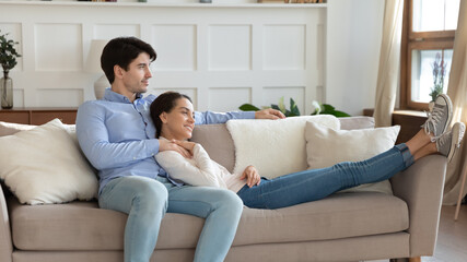Whole life is before. Peaceful loving young spouses newlyweds embracing with warmth and care on sofa at new rented purchased house flat looking aside dreaming pondering imagining happy future together