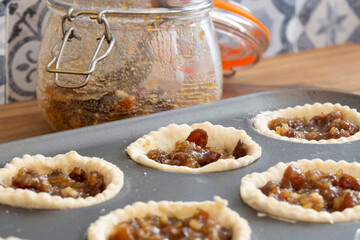 Mince pies in a baking tray with mincemeat in a clip glass jar and a spoon.  On a wood worktop with a tiled background