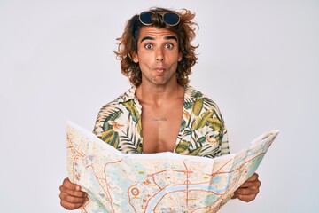 Young hispanic man wearing summer style holding map making fish face with mouth and squinting eyes, crazy and comical.