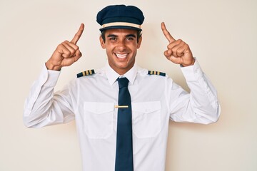 Young hispanic man wearing airplane pilot uniform smiling amazed and surprised and pointing up with fingers and raised arms.