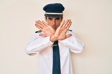 Young hispanic man wearing airplane pilot uniform rejection expression crossing arms and palms doing negative sign, angry face