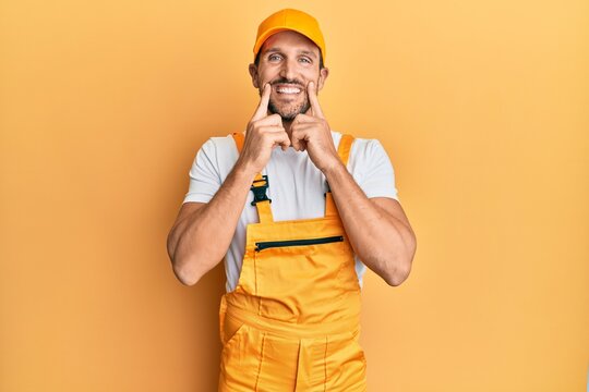 Young handsome man wearing handyman uniform over yellow background smiling with open mouth, fingers pointing and forcing cheerful smile