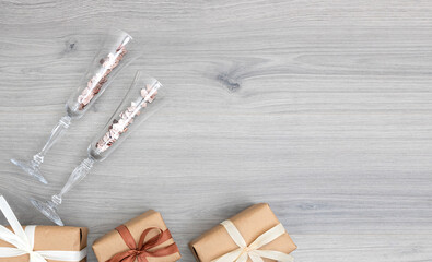 Two champagne flutes with golden confetti and wrapped presents with ribbons on grey wooden background