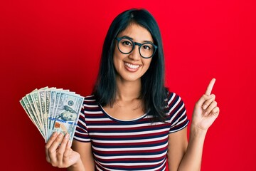 Beautiful asian young woman holding dollars smiling happy pointing with hand and finger to the side