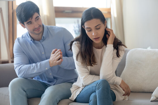 I am so sorry. Guilty millennial man husband partner of young woman sitting on sofa behind her back with clasped hands asking forgiveness excusing expressing wish to reconcile after quarrel conflict