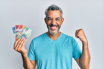 Middle age grey-haired man holding australian dollars screaming proud, celebrating victory and...