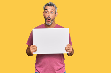 Middle age grey-haired man holding blank empty banner scared and amazed with open mouth for...
