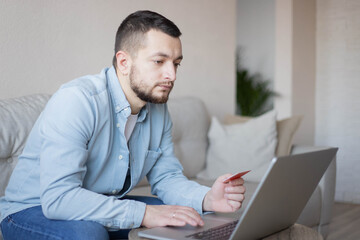 technology, people and online shopping concept - man with laptop and credit card at home.Cheerful guy using credit card and laptop providing online payment sitting on couch at home.