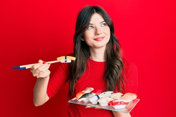 Young beautiful caucasian girl eating sushi using chopsticks smiling looking to the side and staring away thinking.
