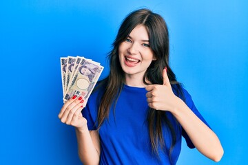 Young beautiful caucasian girl holding 5000 japanese yen banknotes smiling happy and positive, thumb up doing excellent and approval sign