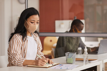 Portrait of modern Asian businesswoman wearing mask and writing in planner while working in office cubicle, copy space