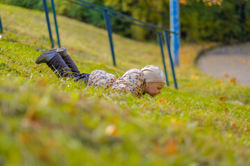 A toddler girl in a jacket and hat lies on her stomach on a hillside. Fun games in the autumn park. Sunny day. Yellow fallen leaves on the grass.