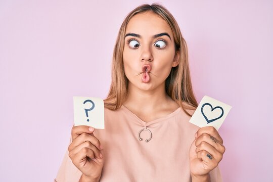 Young beautiful blonde woman holding heart and question mark reminder making fish face with mouth and squinting eyes, crazy and comical.