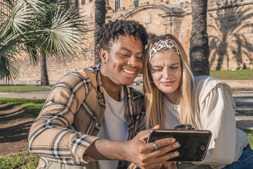 black man with blonde girl sitting on the grass having a lifestyle taking a selfie
