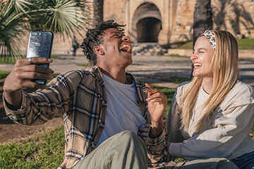 black man with blonde girl sitting on the grass having a lifestyle taking a selfie