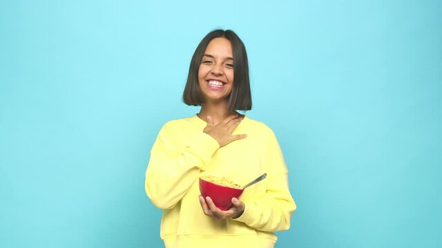 Young latin woman eating a cereals bowl laughs out loudly keeping hand on chest