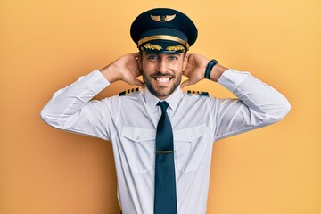 Handsome hispanic man wearing airplane pilot uniform relaxing and stretching, arms and hands behind head and neck smiling happy