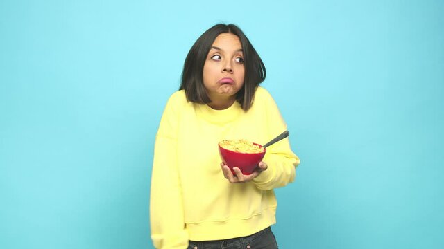 Young latin woman eating a cereals bowl shrugs shoulders and open eyes confused