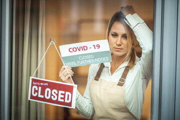 A worried young Caucasian woman standing behind a glass door with the sign ''closed'', holding...