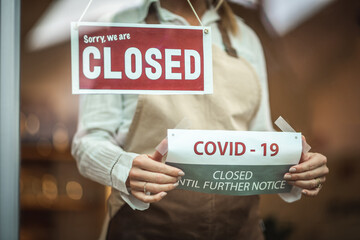 Female silhouette with a beige apron, standing behind the glass door with the sign ''closed'' and putting the covid19 notice
