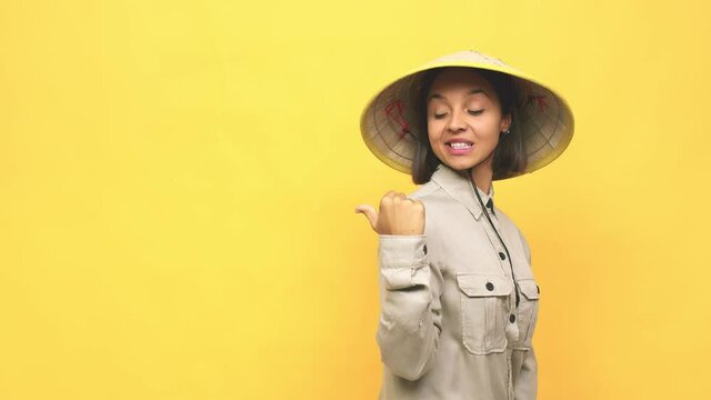 Young latin woman with vietnamite hat  points with thumb finger away, laughing and carefree