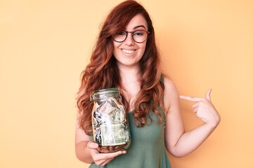 Young beautiful woman wearing glasses holding jar with savings pointing finger to one self smiling happy and proud