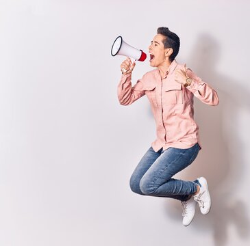 Young beautiful caucasian woman screaming using megaphone. Jumping with thumb up doing ok sign over isolated white background