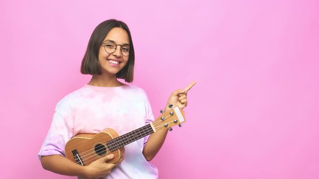 Young latin woman playing ukelele smiling and pointing aside, showing something at blank space