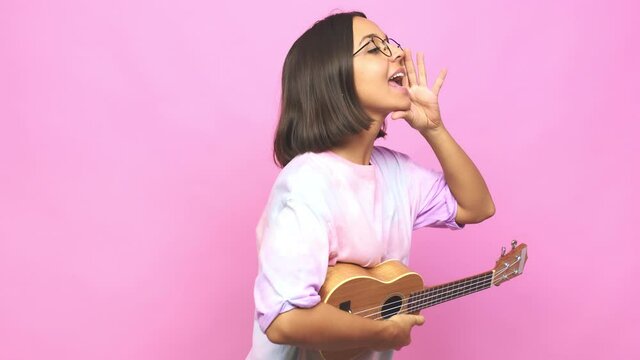 Young latin woman playing ukelele shouting and holding palm near opened mouth