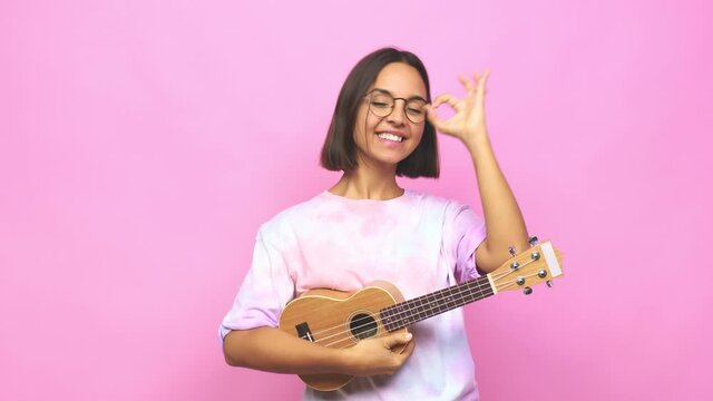 Young latin woman playing ukelele excited keeping ok gesture on eye