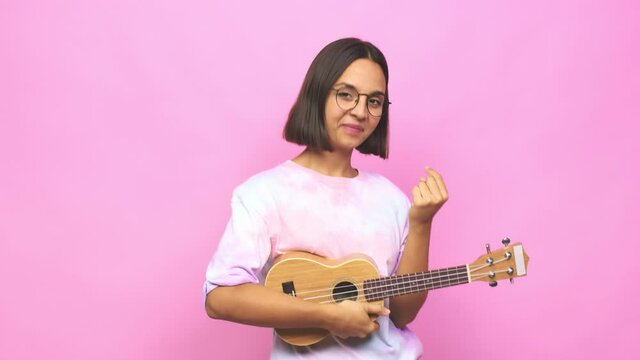 Young latin woman playing ukelele pointing with finger at you as if inviting come closer