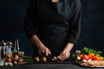 Chef cuts with knife champignons on the table with variety ingredients for cooking mushroom pie on dark blue background. Concept of cooking process. Frozen motion.