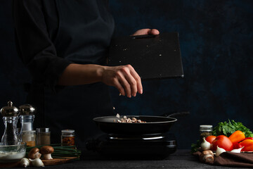 Chef pours pours chopped mushrooms into pan on the table with variety ingredients for cooking pie on dark blue background. Concept of cooking process. Frozen motion. Recipe from cookbook.