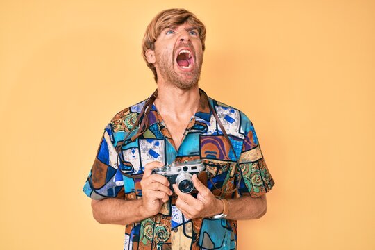 Young blond man holding vintage camera angry and mad screaming frustrated and furious, shouting with anger looking up.