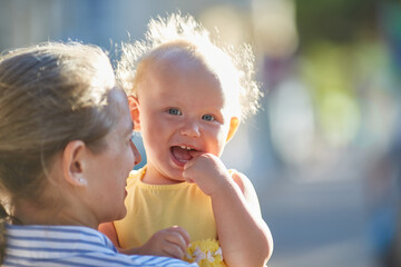 Fototapeta premium Happy smiling baby in mother's arms. Beautiful sunbeams on the street on a summer day.