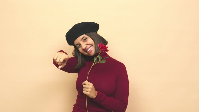 Young latin woman with boina holding rose laughing pointing forward