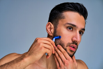 Closeup man with black hair with a beard shaving with blue shaver his cheek. Beauty concept