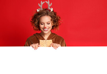 cute funny young woman in a Christmas reindeer costume with blank placard smiles and rejoices on colorful red background.
