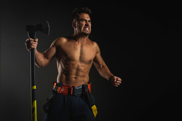 Fototapeta na wymiar young handsome adult, muscular firefighter in uniform holding ax fire equipment in hands, pensive, isolated on dark background. Low key