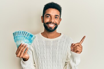 Handsome young hispanic man holding 100 brazilian real banknotes smiling happy and positive, thumb...