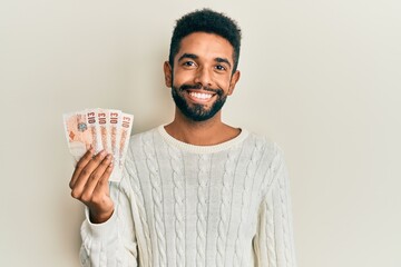 Handsome young hispanic man holding 10 united kingdom pounds banknotes looking positive and happy...