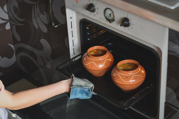 Woman taking out brown ceramic pots with baked or stewed food out of oven. Low DOF. Selective focus.