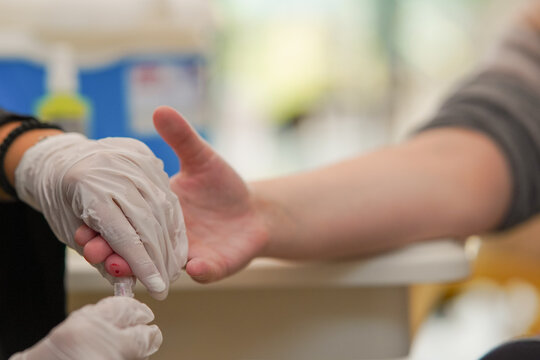 Wider image of a medical nurse taking a blood sample from a finger of a patient into the vial while wearing a sterile gloves in the laboratory