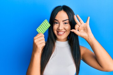 Young beautiful hispanic girl holding birth control pills doing ok sign with fingers, smiling friendly gesturing excellent symbol