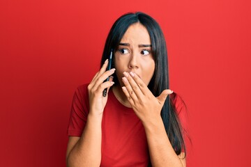 Young beautiful hispanic girl having conversation talking on the smartphone covering mouth with...