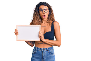 Young hispanic woman with tattoo holding empty white chalkboard scared and amazed with open mouth...